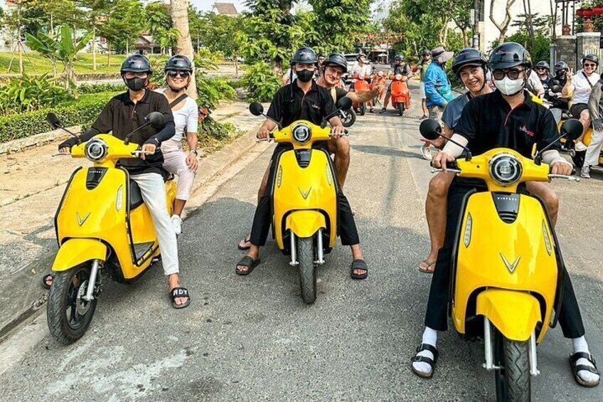 People riding yellow scooters on a road with trees and buildings in the background. 4