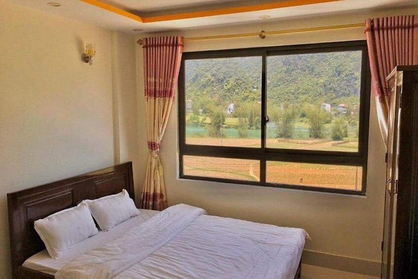 A bedroom with a bed, curtains, a window with a view of greenery, and a wall-mounted light fixture. 4