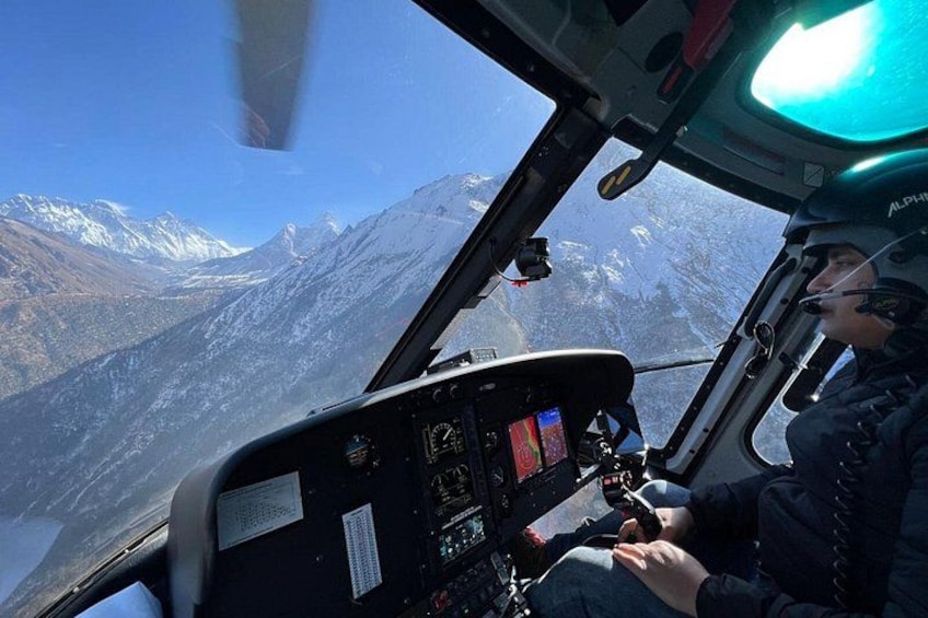 Helikoptercockpit med pilot, kontrolpanel og et bjergrigt landskab i baggrunden. 4