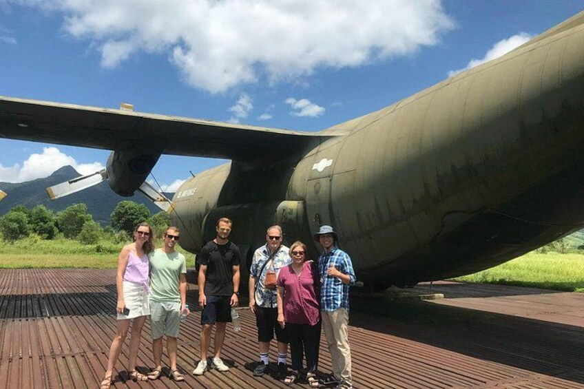 Un grupo de personas de pie frente a una aeronave militar sobre una cubierta de madera, con cielo despejado al fondo. 3