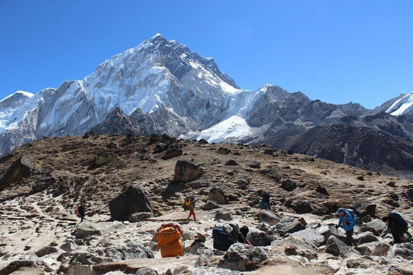 A mountainous landscape with a prominent snow-capped peak, rocky terrain, and hikers with backpacks. 2