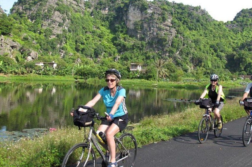Twee fietsers op een verhard pad langs een rustig meer, met weelderige begroeiing en rotsachtige heuvels op de achtergrond. 5