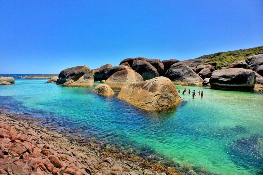 Ett kustlandskap med stora stenblock i klart turkost vatten, en stenig strand och en klarblå himmel. 4