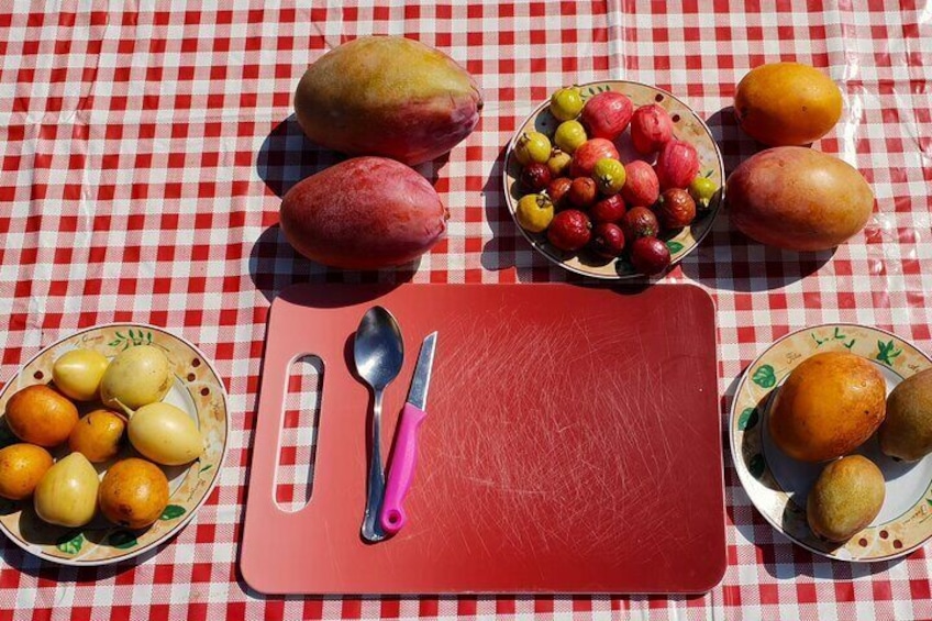 A table with a red cutting board, two spoons, and bowls of fruits including mangoes, apples, and oranges. 3