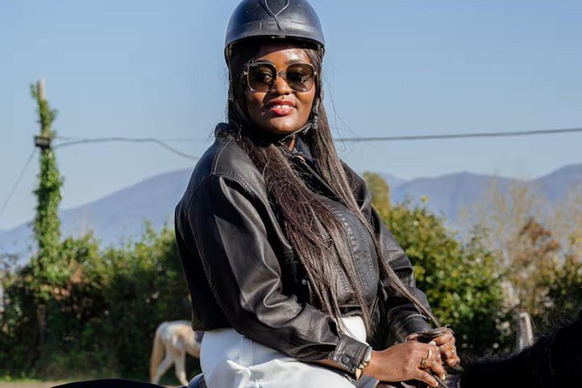Una persona con chaqueta de cuero negra y casco, de pie al aire libre, con follaje y montañas de fondo. 2