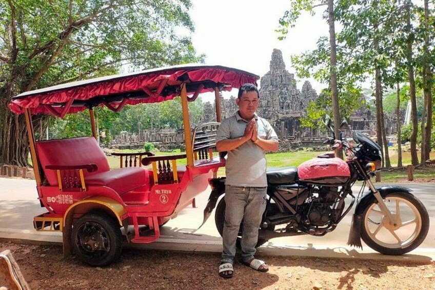 A man standing next to a red rickshaw and a motorcycle with a temple in the background. 4