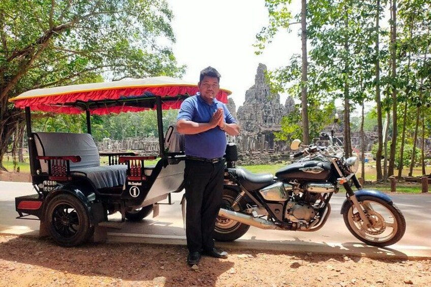 A man standing next to a motorcycle and a three-wheeled vehicle with a red canopy, with a temple in the background. 5