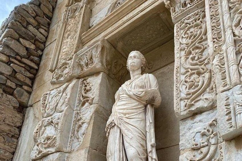 A stone statue of a figure with draped clothing, set against a detailed architectural backdrop with carved patterns and a stone wall. 3