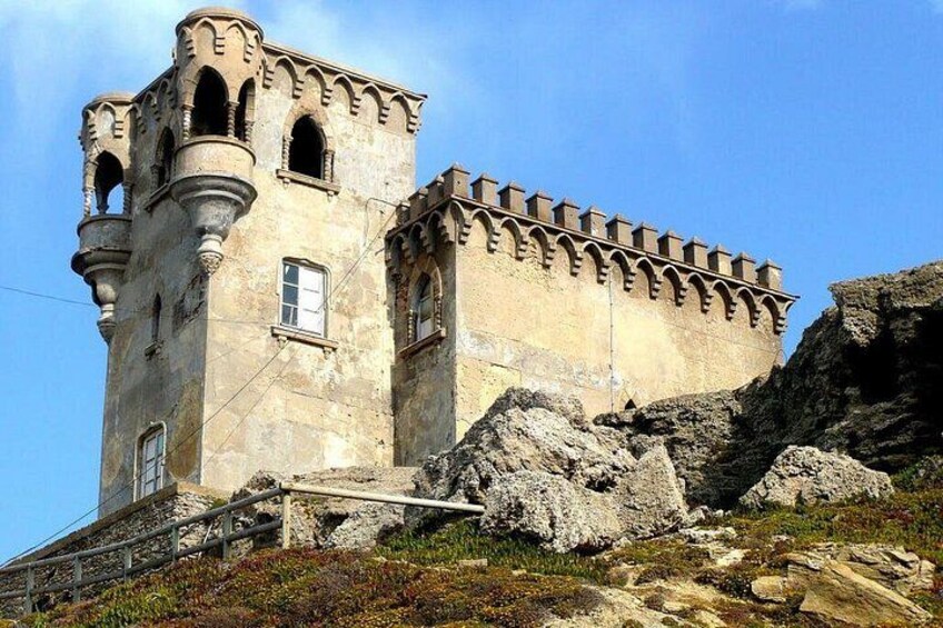 A historic stone tower with arched windows and a fortified wall, situated on a rocky hillside. 2