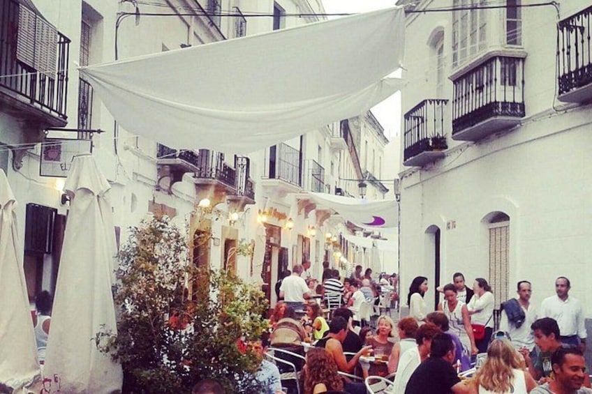 A crowded outdoor café with white umbrellas and tables, surrounded by buildings with balconies. 3