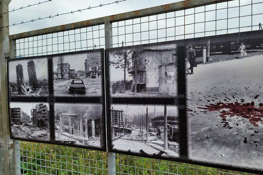 A series of black and white photographs on a fence, depicting urban scenes with buildings and vehicles. 3