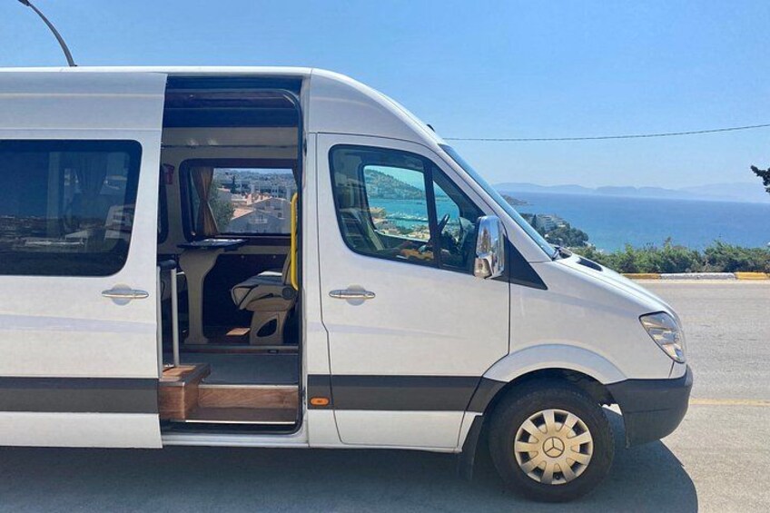 A white van parked on a road with a clear view of the sea and distant hills in the background. 1