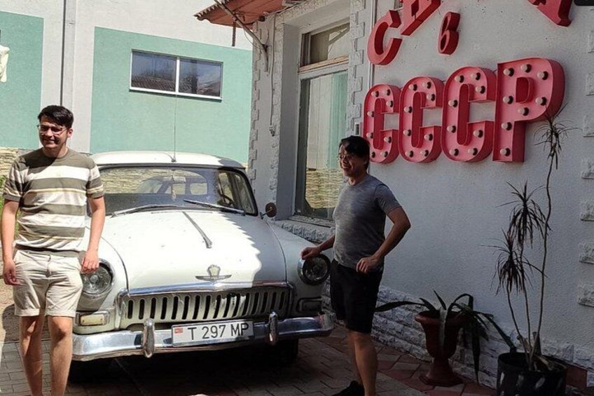 A vintage car parked in front of a building with a large red sign that reads 'CCCP'. 2