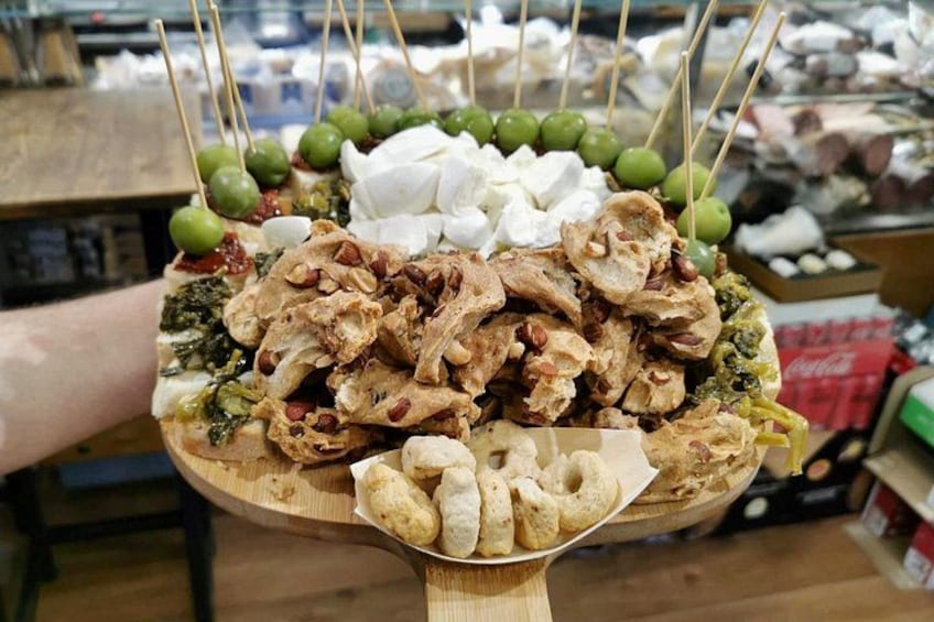 A wooden platter with various food items, including olives, nuts, and cheese, held by a person. 1
