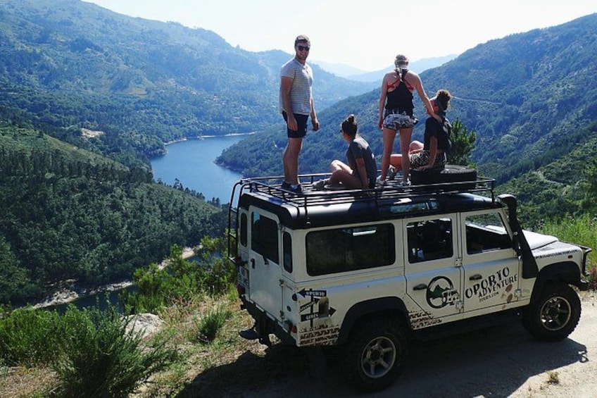 Een witte terreinwagen met 'OZPORTO ADVENTURE' op de zijkant, geparkeerd aan de kant van de weg, met een prachtig uitzicht op een meer en bergen op de achtergrond. 3