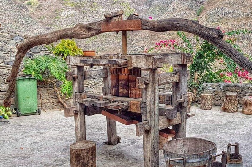 A rustic wooden structure with a large branch arching over it, surrounded by greenery and stone walls. 5