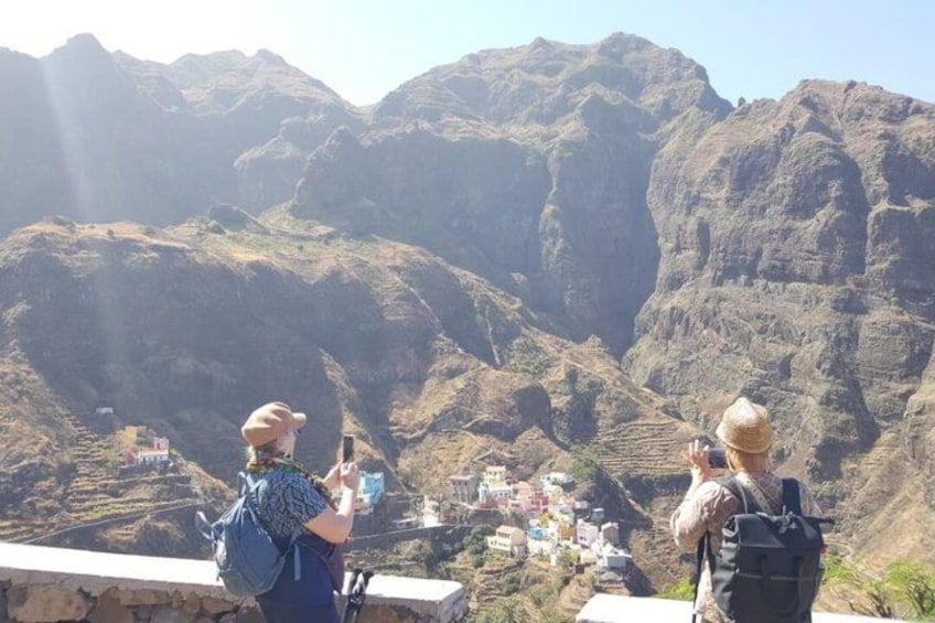 Two people taking photographs of a mountainous landscape with terraced fields and a small village below. 1