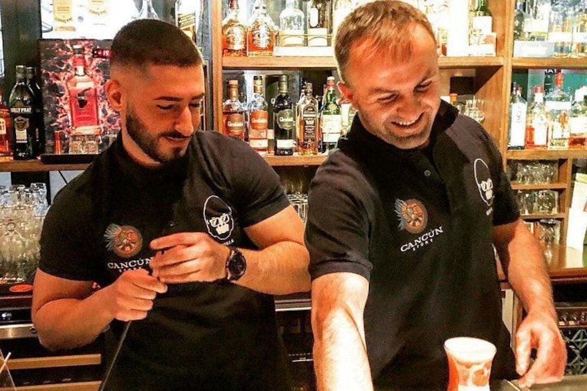 Two people wearing black shirts with a logo, standing in a bar with shelves of bottles and glasses. 2