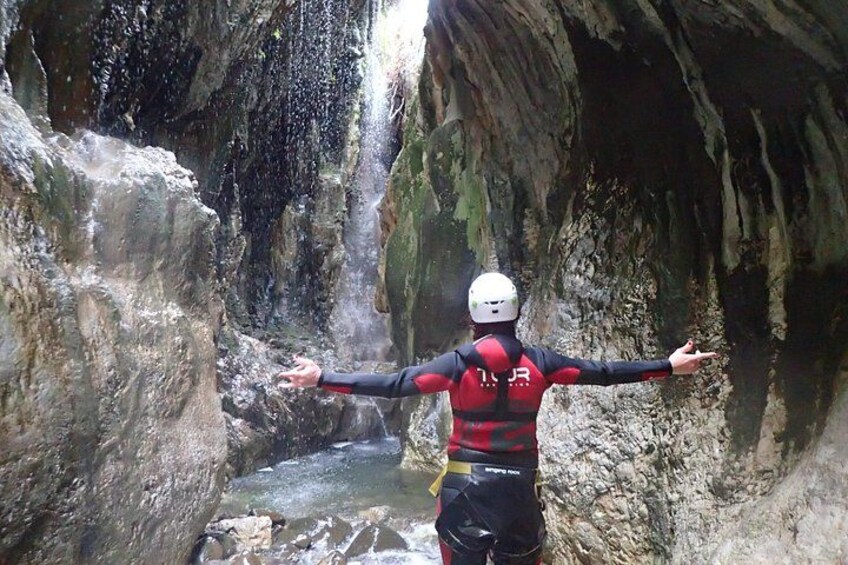 Une personne vêtue d’une combinaison de plongée rouge et noire, portant un casque, se tient dans une grotte rocheuse, les bras tendus, face à une cascade. 4