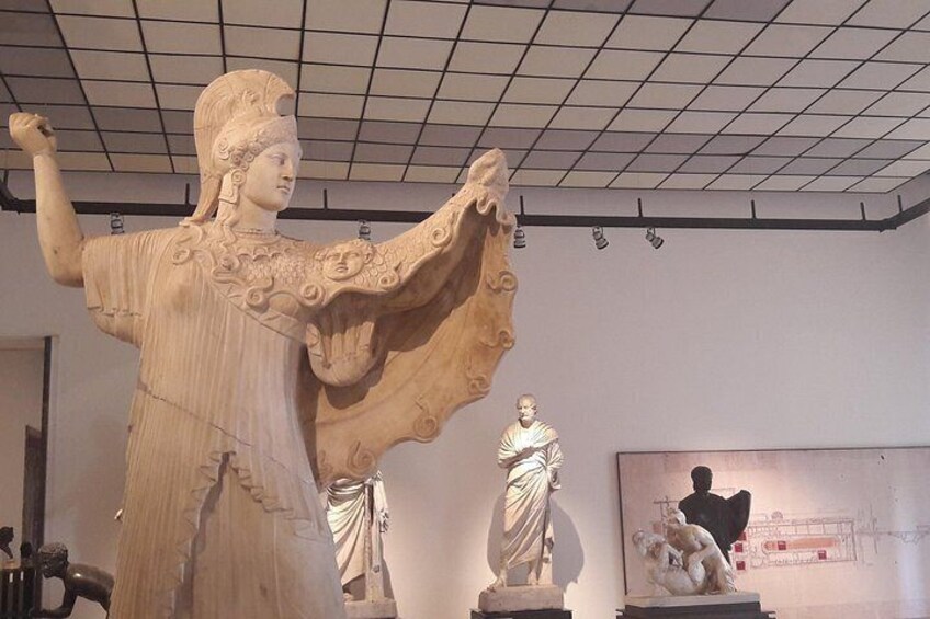 A large classical statue with draped clothing, a smaller statue in the background, and a framed diagram on the wall. 1