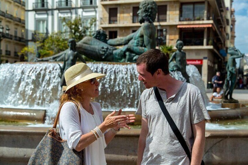 Two people conversing near a fountain with statues in the background. 1