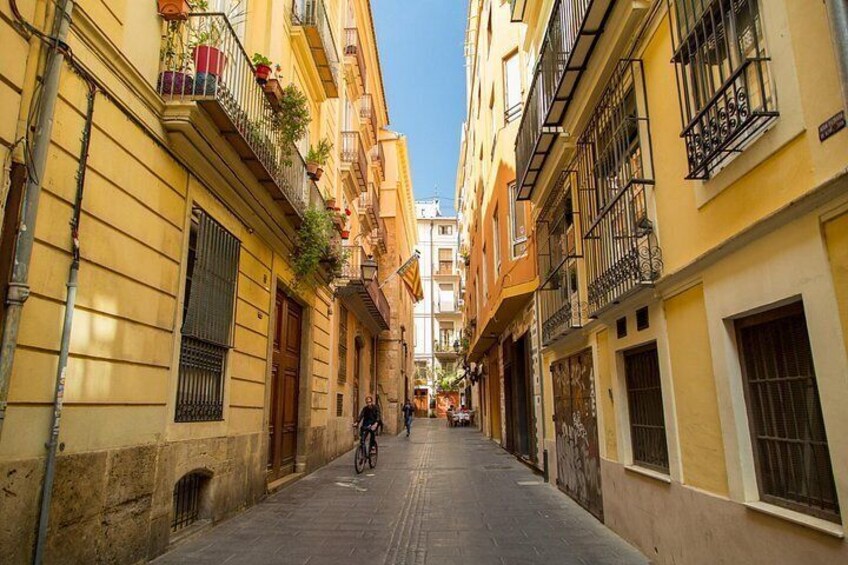 A narrow street lined with yellow buildings, a person riding a bicycle, and a clear blue sky. 5