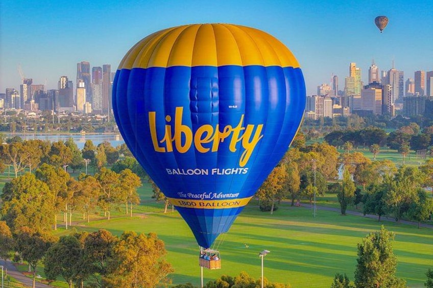 A hot air balloon with 'Liberty Balloon Flights' written on it, flying over a park with a city skyline in the background. 1