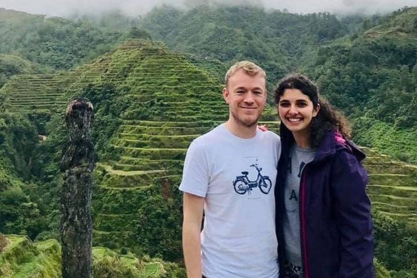 Two people standing in a terraced landscape with lush greenery and a prominent tree trunk. 3