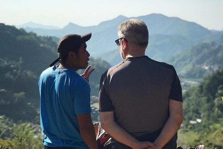 Two people standing and conversing with a mountainous landscape in the background. 3