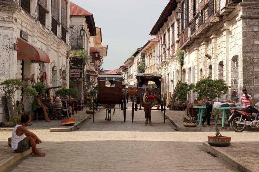A cobblestone street with buildings, a horse-drawn cart, and a person sitting on the ground. 5