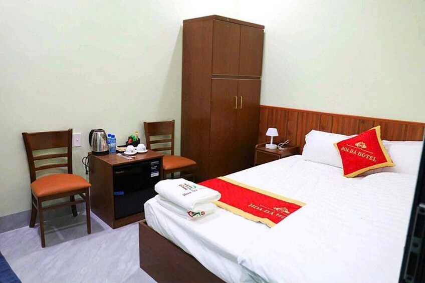 A hotel room with a bed, two wooden chairs, a desk with a kettle, a nightstand, and a wardrobe. 5