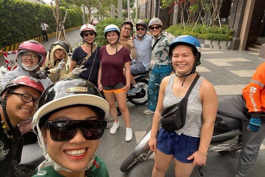 Een groep mensen op scooters, sommigen dragen helmen, in een stedelijke omgeving met gebouwen en bomen. 3