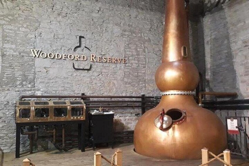 Un grande alambicco in rame, botti di legno e un cartello con su scritto 'Woodford Reserve' in un ambiente rustico. 2
