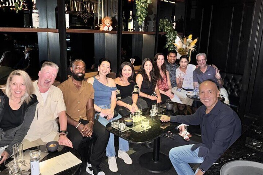 A group of people seated around a table with drinks and a menu, in a dimly lit indoor setting with shelves and plants in the background. 3