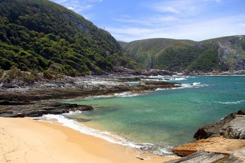 A coastal landscape with a sandy beach, clear turquoise waters, rocky outcrops, and green hills. 2
