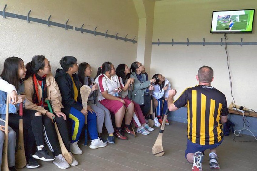 A group of people sitting on chairs, some holding wooden sticks, in a room with a television displaying a sports game. 3