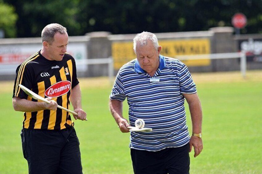 Two men on a grassy field, one holding a stick and the other holding a ball. 4