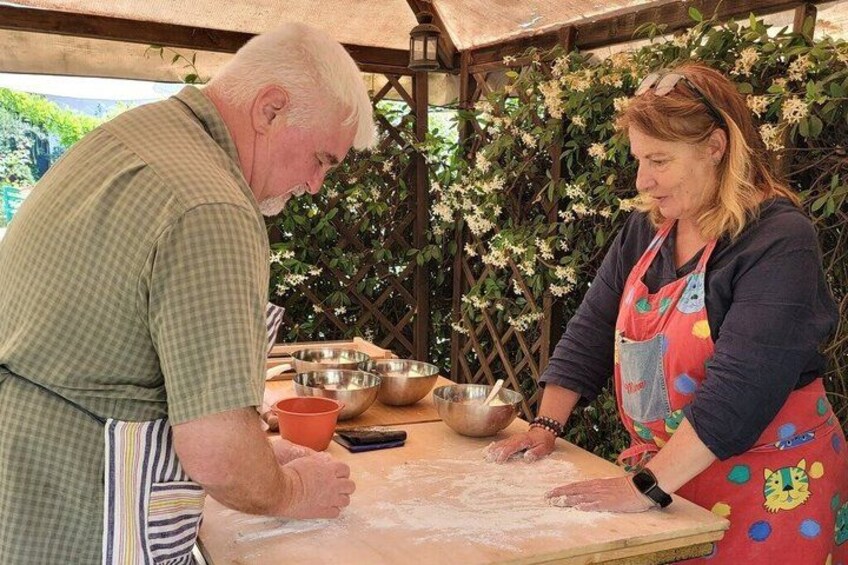 Zwei Personen backen draußen an einem Holztisch, mit Küchenutensilien und einer Gitterstruktur im Hintergrund. 2