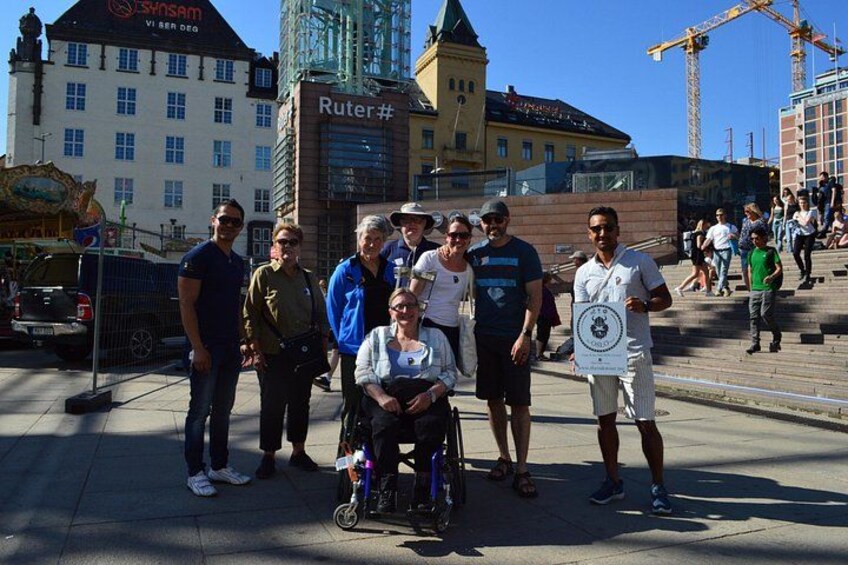 A group of people, including a person in a wheelchair, standing on a paved area with buildings and a crane in the background. 4