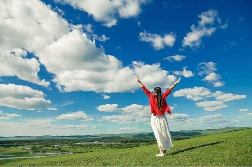 Eine Person steht mit erhobenen Armen auf einer grasbewachsenen Anhöhe, bewölkter blauer Himmel und eine ferne Landschaft im Hintergrund. 2