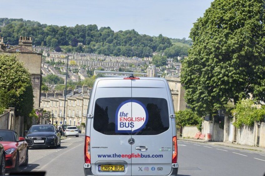 Ein weißer Van mit dem Schriftzug 'The English Bus' und der Webseite auf der Rückseite fährt auf einer Straße, im Hintergrund sind Gebäude und Bäume zu sehen. 2