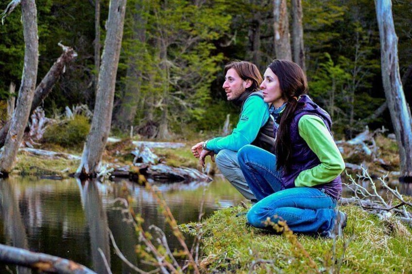 Two people crouching by a calm river in a forested area. 2