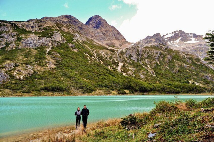 Un paisaje montañoso con un lago turquesa, dos personas de pie en la orilla y un pico montañoso destacado al fondo. 4