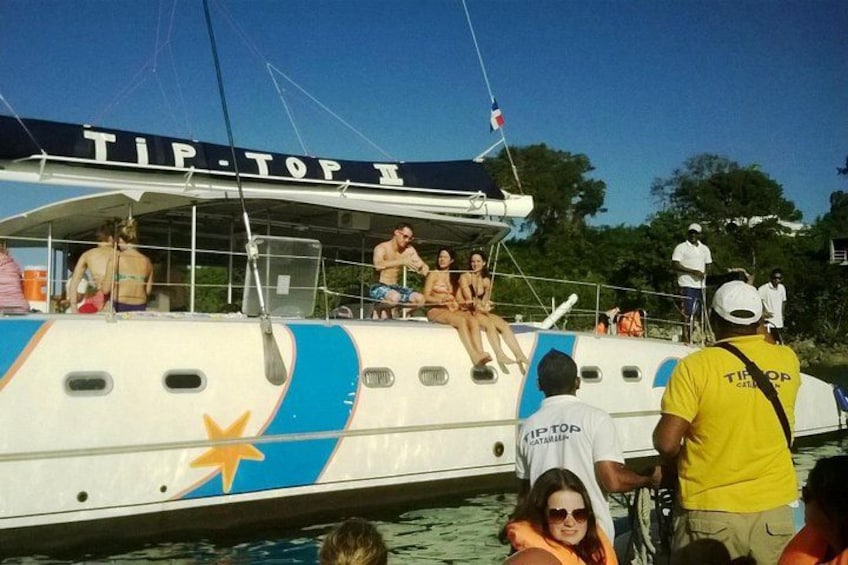 Een boot met mensen op het dek en sommigen in het water, een vlag en een bord met de tekst 'TIP TOP II'. 2