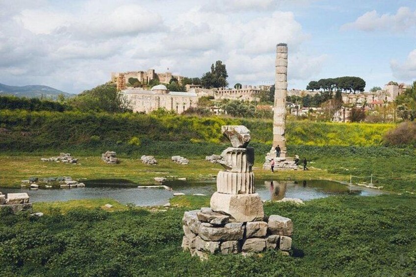 A historical site with ruins, a tall column, and a statue in a grassy area with a pond. 2