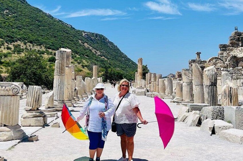 Two people standing in an ancient ruin with columns and a mountainous backdrop. 4
