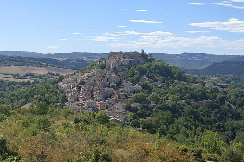 Une ville construite sur une colline, surmontée d’une structure ressemblant à un château, entourée de verdure et avec d’autres collines en arrière-plan. 1