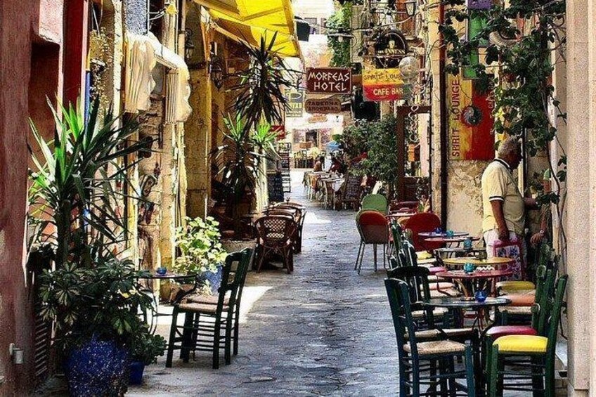 A narrow street with outdoor seating, plants, and signs for a hotel and cafe. 1
