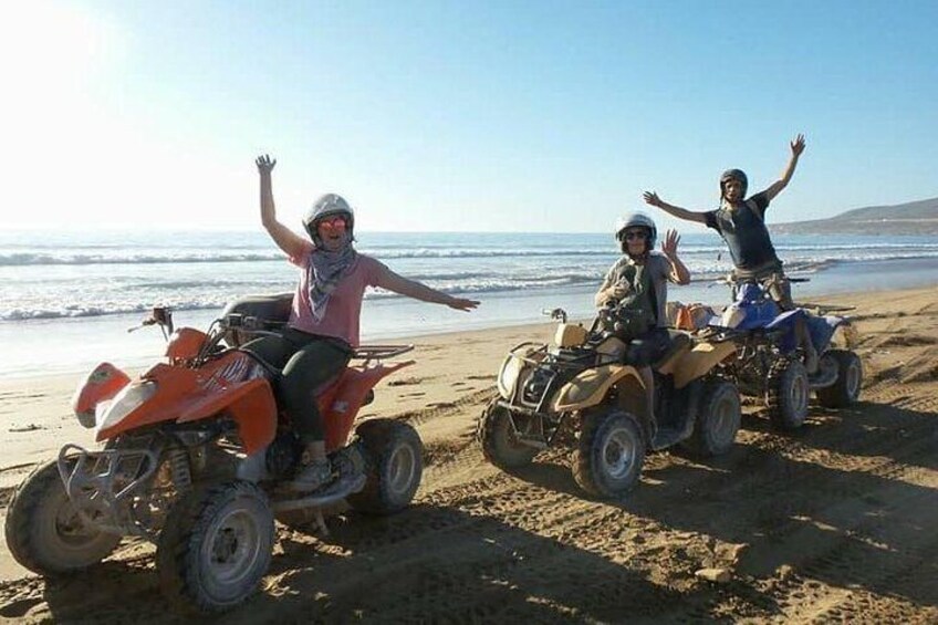 Drie personen op terreinwagens op een zanderig strand, met de oceaan op de achtergrond. 1