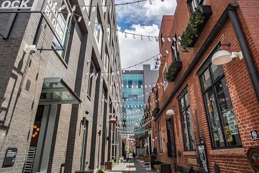 Une rue étroite en milieu urbain, bordée d’immeubles en briques, illuminée de guirlandes lumineuses, avec un bâtiment moderne en verre en arrière-plan. 2